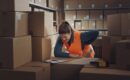 A warehouse worker managing back pain as she works with boxes and a clipboard.