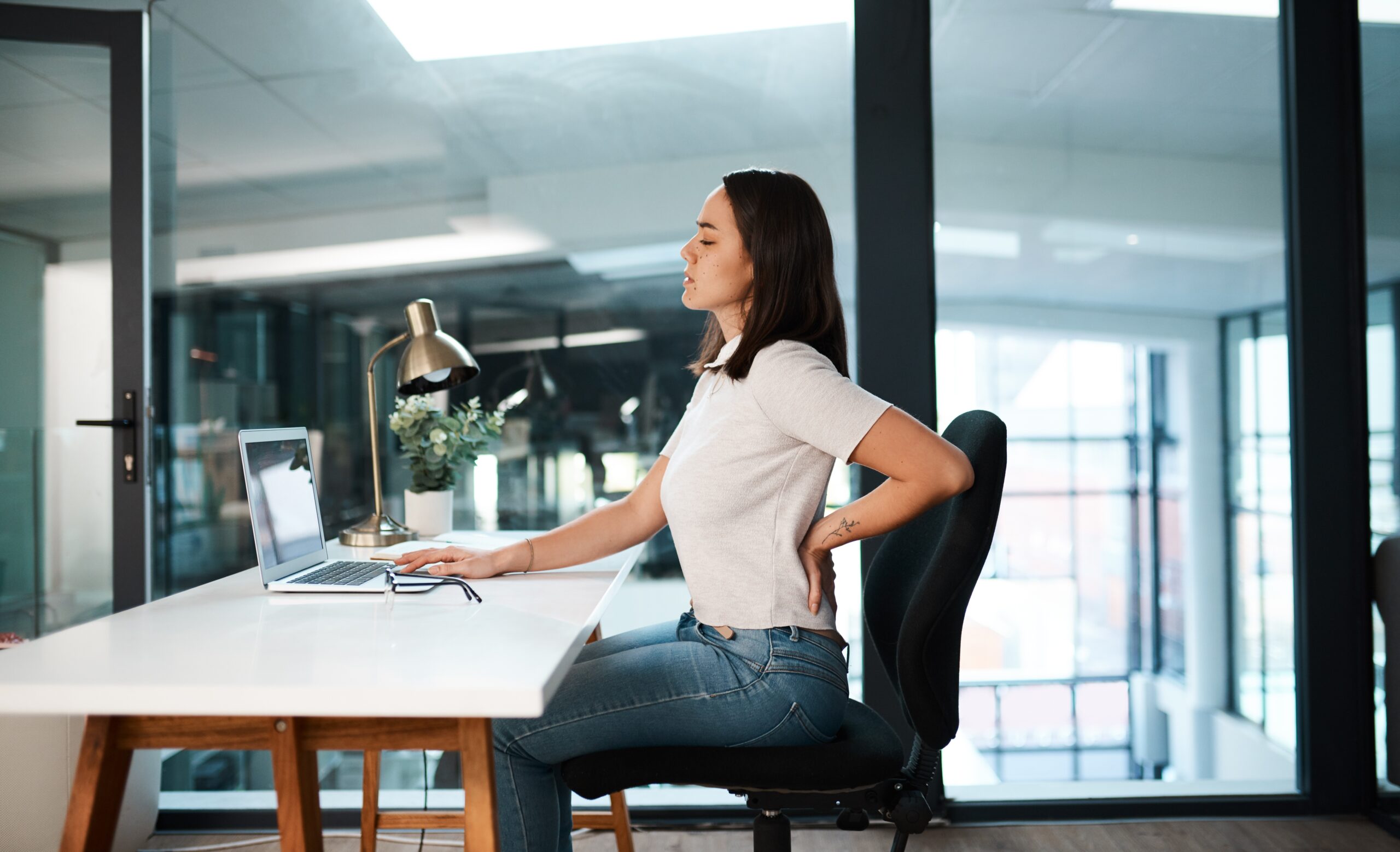 A female office employee sitting at her desk, managing back pain while she tries to work.