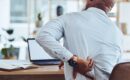 A male office worker rubs his back while working at a computer office desk and managing back pain the best he can.