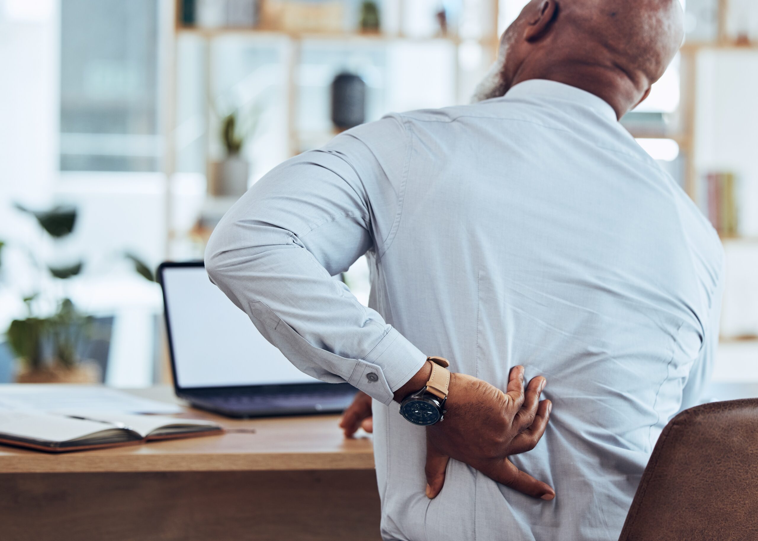 A male office worker rubs his back while working at a computer office desk and managing back pain the best he can.